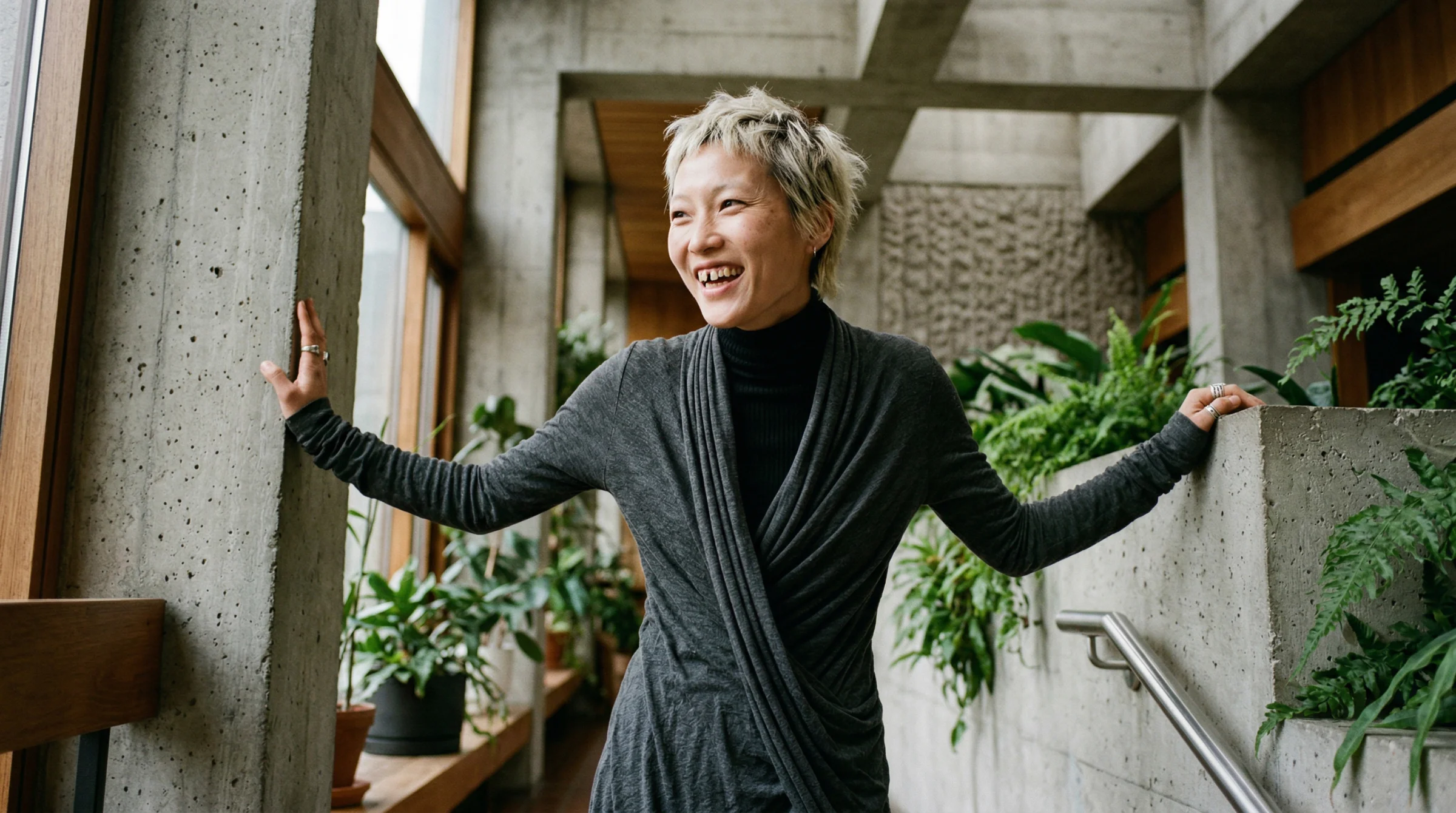The benchmark shot. East Asian woman, late 20s, gap teeth and cropped bleached hair, laughing candidly, framed by plants and a concrete railing, shot at 24mm on a Leica Q3. This is what "four tokens plus framing rules plus deep chiaroscuro plus 24mm" produces when everything clicks.